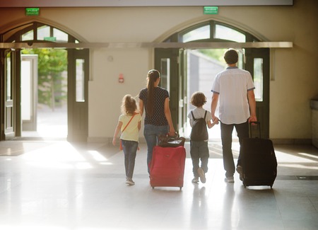 Young family in the waiting room at the station. Dad holds the hand of her son, mother-daughter. Also parents roll big suitcases. View from the back. Sun through large windows illuminate the hall.の写真素材