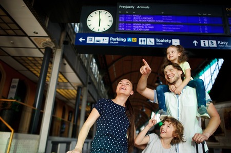 Cheerful young family at the train station. Father, mother, son and daughter are under an electronic scoreboard. Father holding small daughter on his shoulders. Everyone looks wherever shows father.の写真素材