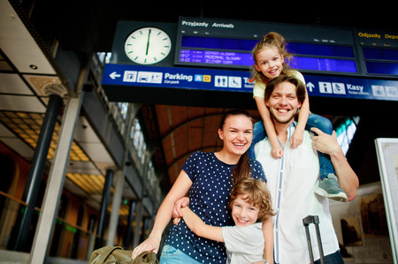 Young couple with two cute kids at the station. Man holding daughter on his shoulders and hugging his wife and son. All smiling happily. Above them, an electronic scoreboard with a schedule.の写真素材