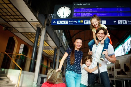 Young couple with two children at the station.Man holding daughter on shoulders. Son hugging dad. Woman holds the hand luggage. All smiling happily. Above them an electronic scoreboard with a scheduleの写真素材
