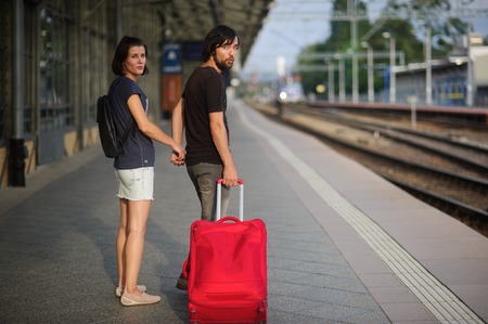 Young couple standing on the platform holding hands. Young man rolls the red suitcase. Both looking into the camera.の写真素材