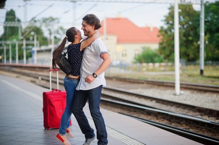 On the empty platform joyful meeting. The young man lifted the girl. Couple hugging. Young people laugh gleefully. Nearby is the red suitcase.の写真素材