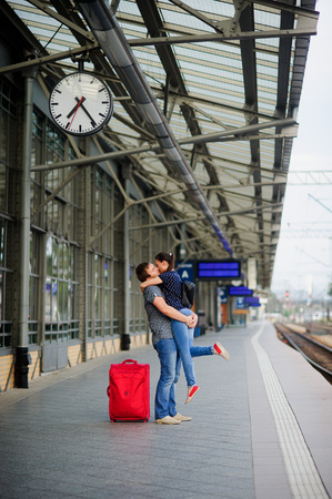 Joyful meeting under the clock at the railway station. The young man lifted the girl. Couple embraces and kisses. Young people with tenderness look at each other. Nearby is the red suitcase.の写真素材