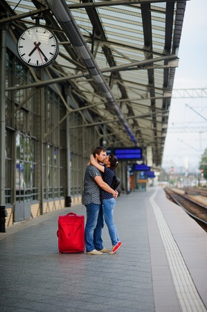 Joyful meeting under the clock at the railway station. The young man lifted the girl. Couple hugging and kissing. Nearby is the red bag.の写真素材