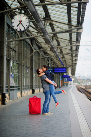 Joyful meeting under the clock at the railway station. The young man lifted the girl. Young people with tenderness look at each other. Nearby is the red suitcase.の写真素材