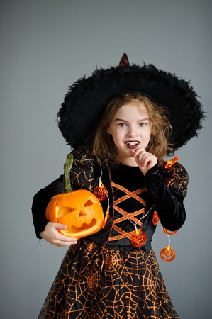 Halloween. Girl portray evil sorceress.She is wearing black-and-orange dress and hat. From under his hat sticking disheveled hair. Girl has an evil expression. In hands at her pumpkin - Jack o lanternの写真素材