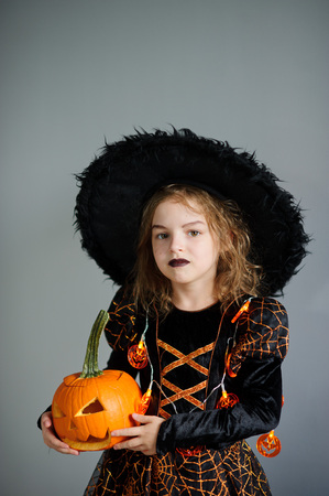Halloween. Girl portray evil sorceress.She is wearing black-and-orange dress and hat. From under his hat sticking disheveled hair. Girl has an evil expression. In hands at her pumpkin - Jack o lanternの写真素材