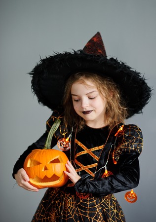 Portrait of the girl of 8-9 years in a suit for Halloween. She represents the evel sorcerer. The girl is dressed in a black-orange dress a hat. In hands at her pumpkin - Halloween symbol.の写真素材