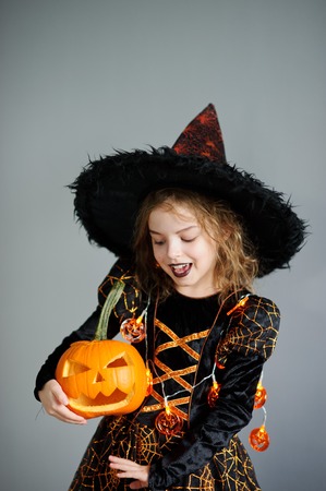 The girl in a costume for Halloween. She portray the evil fairy. On the girl a black-orange dress and a big hat. From under his hat sticking disheveled hair. In hands at her pumpkin - Halloween symbol.の写真素材