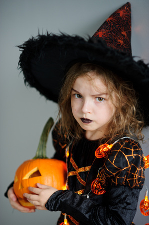 Portrait of the girl of 8-9 years in a suit for Halloween. She represents the evel sorcerer. The girl is dressed in a black-orange dress a hat. In hands at her pumpkin - Halloween symbol.の写真素材