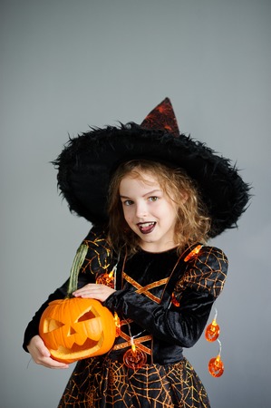 Portrait of the girl of 8-9 years in a suit for Halloween. She represents the evel sorcerer. The girl is dressed in a black-orange dress a hat. In hands at her pumpkin - Halloween symbol.の写真素材
