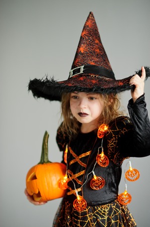 Portrait of the girl of 8-9 years in a suit for Halloween. She represents the evel sorcerer. The girl is dressed in a black-orange dress a hat. In hands at her pumpkin - Halloween symbol.の写真素材