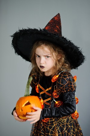 Halloween. Girl portray evil sorceress.She is wearing black-and-orange dress and hat. From under his hat sticking disheveled hair. Girl has an evil expression. In hands at her pumpkin - Jack o lanternの写真素材