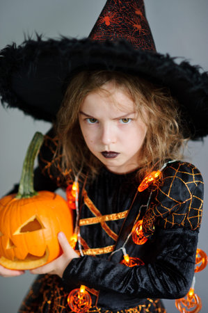 Halloween. Girl portray evil sorceress.She is wearing black-and-orange dress and hat. From under his...