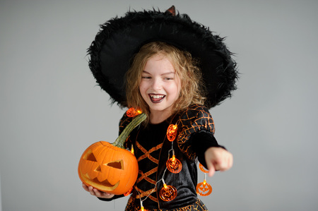 Portrait of the girl of 8-9 years in a suit for Halloween. She represents the evel sorcerer. The girl is dressed in a black-orange dress a hat. In hands at her pumpkin - Halloween symbol.の写真素材