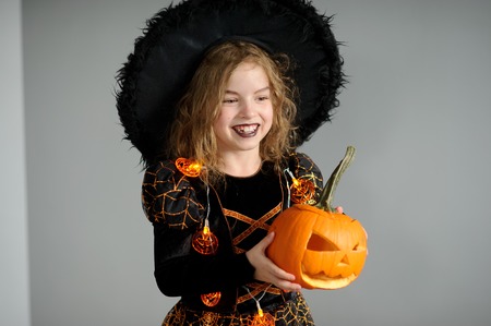Portrait of the girl in a suit for Halloween. She represents the evel sorcerer. The girl is dressed in a black-orange dress a hat. In hands at her pumpkin - Halloween symbol. Children like Halloween.の写真素材