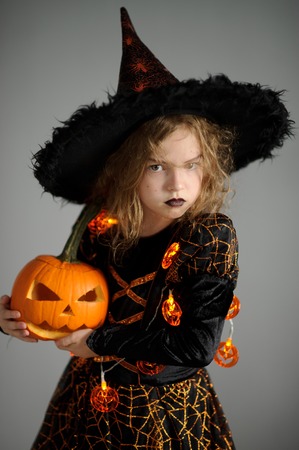 Halloween. Girl portray evil sorceress.She is wearing black-and-orange dress and hat. From under his hat sticking disheveled hair. Girl has an evil expression. In hands at her pumpkin - Jack o lanternの写真素材