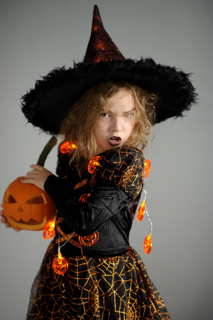 Halloween. Girl portray evil sorceress. She is wearing a black-and-orange dress and hat. From under his hat sticking disheveled hair. Girl has an evil expression. In hands at her pumpkin - Jack o lanternの写真素材