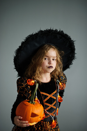 Portrait of the girl of 8-9 years in a suit for Halloween. She represents the evel sorcerer. The girl is dressed in a black-orange dress a hat. In hands at her pumpkin - Halloween symbol.の写真素材