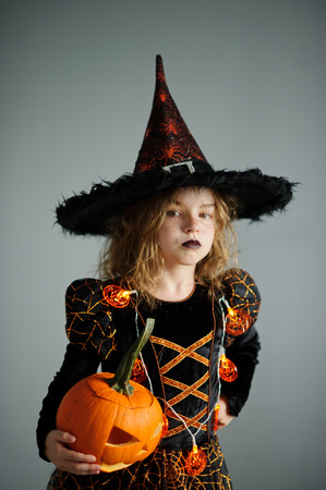 Halloween. Girl portray evil sorceress. She is wearing black-and-orange dress and hat.From under his hat sticking disheveled hair. Girl has an evil expression. In hands at her pumpkin - Jack o lanternの写真素材