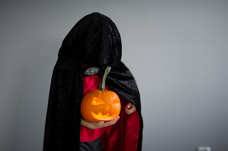 Child is dressed in black -red toga with hood. It is a costume for Halloween. He represents the mysterious wizard. The hood covers the face. Child holds pumpkin with a candle inside - Halloween symbol.の写真素材