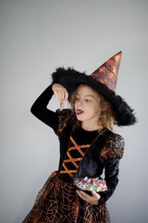 Halloween Party. Girl 8-9 years in image the evil sorcerer. On the girl a black-orange dress and a big hat. Girl holds sweets in hands. She has a ridiculous look. Trick or Treat.の写真素材
