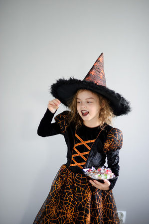 Halloween Party. Girl 8-9 years in image the evil sorcerer. On the girl a black-orange dress and a big hat. Girl holds sweets in hands. She has a ridiculous look. Trick or Treat.の写真素材