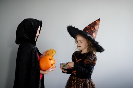 Boy and girl in suits for Halloween. They in image powers of darkness. Girl - evil fairy, boy - warlock. Girl holds sweets in hands. Boy holds pumpkin with a candle inside - Halloween symbol. Booの写真素材