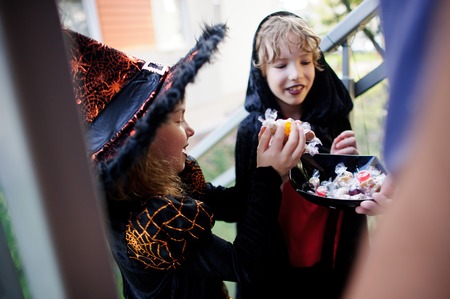 Two children, boy and girl, in dark costumes for Halloween take candies. In the foreground a hand with the vase filled with sweets.Trick or Treat. Children love Halloween.の写真素材