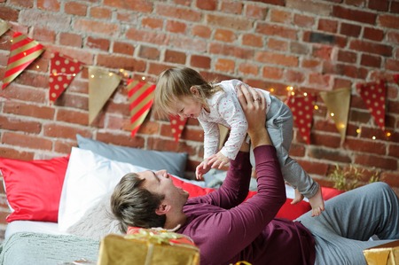 Christmas morning. Young man cheerfully plays with small daughter. On a bed there are many Christmas gifts in bright packings. The room is festively decorated. Merry Christmas and a Happy New Year.の写真素材