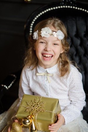 Beautiful girl photographed for Christmas card. Girl is dressed a white-gold dress with a wreath on her head. She looks like a Christmas angel. In hands of the girl the Christmas gift in gold packing.の写真素材