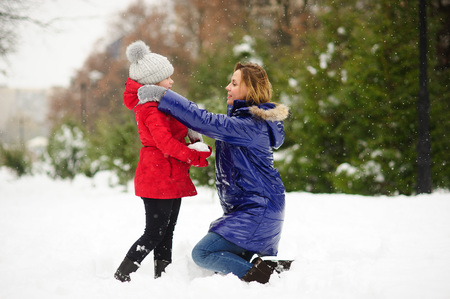Young woman and her daughter builds snowman. Woman and girl are dressed in bright and warm jackets. It is snowing. Mom lovingly corrects daughters jacket. All earth is covered with fluffy snow.の写真素材