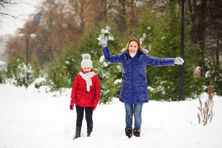 Young woman with the daughter on walk in winter day. Woman and the girl are dressed in bright down-padded coats. Woman without headdress. It is snowing. All ground in the park is covered with snow.の写真素材