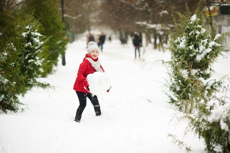 Cheerful girl of younger school age builds snowman. She holds a huge snow sphere in hand and laughs. It is snowing. All ground in park is covered with snow. Girl in bright red jacket and knitted hat.の写真素材