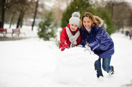 Mother with daughter of younger school age build a snowman in the yard. They have rolled a huge lump of snow. It is heavy, but it is cheerful. It is snowing. Ground is covered with fluffy snowdrifts.の写真素材