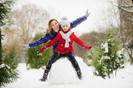 Young woman and girl of school age build a snowman. It is snowing. Ground in park is covered with snow. Woman and girl are dressed in bright down-padded coats. They rejoice to snow and have fun.の写真素材