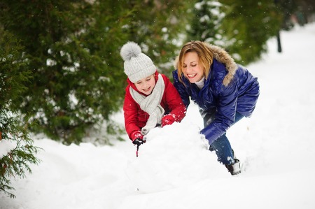 Mother with daughter build a snowman in park. It is snowing. All ground in park is completely covered with fluffy snow. Woman and girl are dressed in bright down-padded coats. They have joyful faces.の写真素材