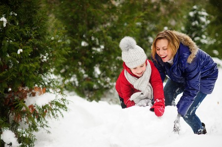 Young woman and girl of school age build a snowman. All ground in park is completely covered with fluffy snow. Woman and girl are dressed in bright down-padded coats. They have a good mood.の写真素材