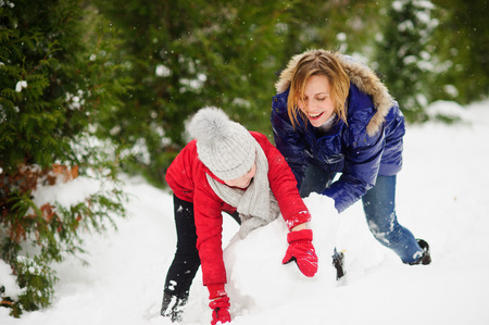 Young woman and girl of school age build a snowman. All ground in park is completely covered with fluffy snow. Woman and girl are dressed in bright down-padded coats. Active recreation, useful to health.の写真素材