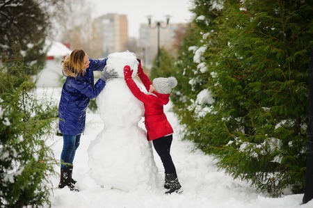 The young woman and her daughter build a snowman. They establish the head to a big snowman. In the park it is snowing, snowdrifts lie. Mother and the daughter cheerfully spend time.の写真素材