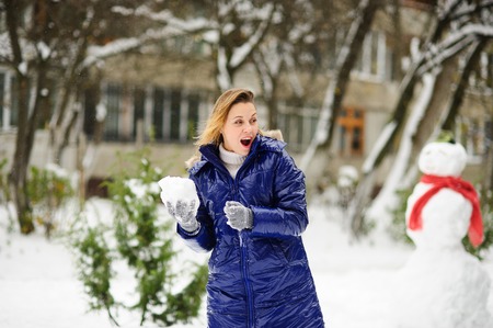 Young woman plays snowballs. It is snowing. Earth is covered with snowdrifts. Woman is dressed in blue warm jacket, without headdress. Excited by play, she does not feel cold. Nearby there is snowman.の写真素材