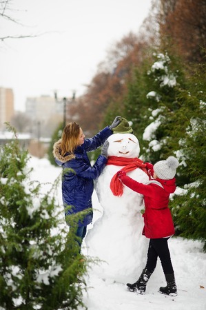 Mother with daughter of school age have built a snowman. Woman putting on his hat on the snowman. Girl tying his scarf. All ground in park is completely covered with fluffy snow.の写真素材