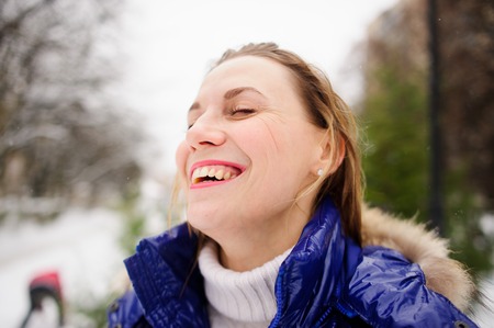 Portrait of young woman against the background a winter landscape. She is dressed in bright blue down-padded coat, without headdress. Snowflakes on her hair. Woman has closed eyes and laughs.の写真素材