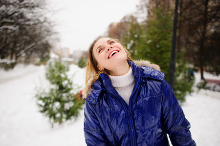 Portrait of young woman against the background a winter landscape. She is dressed in bright blue down-padded coat, without headdress. Snowflakes on her hair. Woman has closed eyes and laughs.の写真素材