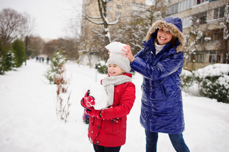 Young woman and her daughter cheerfully spend time on street in winter day. They are dressed in warm jackets. Ground is covered with snow. Mom puts on head to daughter a big lump of snow. Girl laughs.の写真素材