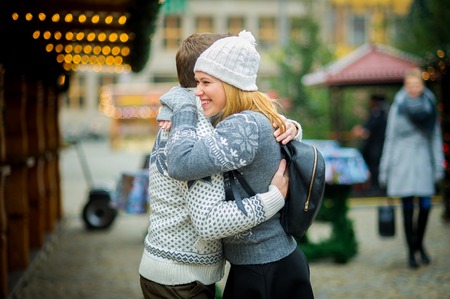 Eve of Christmas. Cute young couple has a good time at the Christmas bazaar. Young people stand in the middle of the fairground and joyfully embrace. Stall is festively decorated with shining garlands.の写真素材