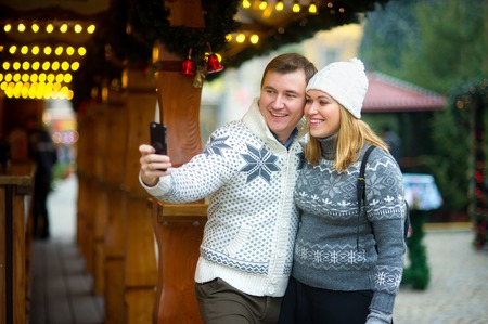 Christmas bazaar. The young couple is photographed near the wooden festively decorated pavilion. A man holds a smart phone in your hand and makes a selfie. Both with pleasure pose. Merry Christmas.の写真素材