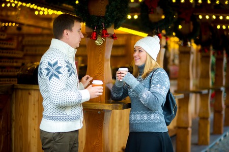 Eve of Christmas. Cute young couple has a good time at the Christmas bazaar. Young people stand near a wooden counter with paper cups in hands. Stall is festively decorated with wreaths and garlands.の写真素材