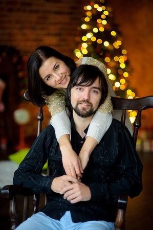 Christmas evening. Young woman embraces man sitting in a chair. In a room corner stands Christmas tree. She shines of fairy lights. Young people gently hold each other by hands. They look in camera.の写真素材