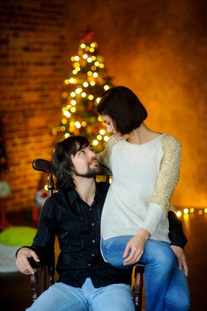 Christmas evening. Young woman embraces man sitting in a chair. She sits on an armrest. Young people with love look at each other. In a room corner stands Christmas tree. She shines in the twilight.の写真素材